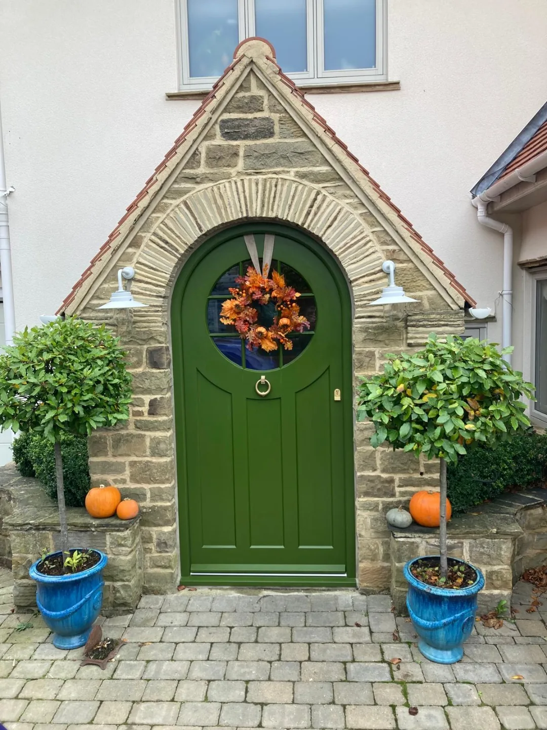 Racing green Gothic arched front door with stone gable arch, brass ring knocker and matching bay topiaries — bespoke heritage door by Bailey Hague Joinery.