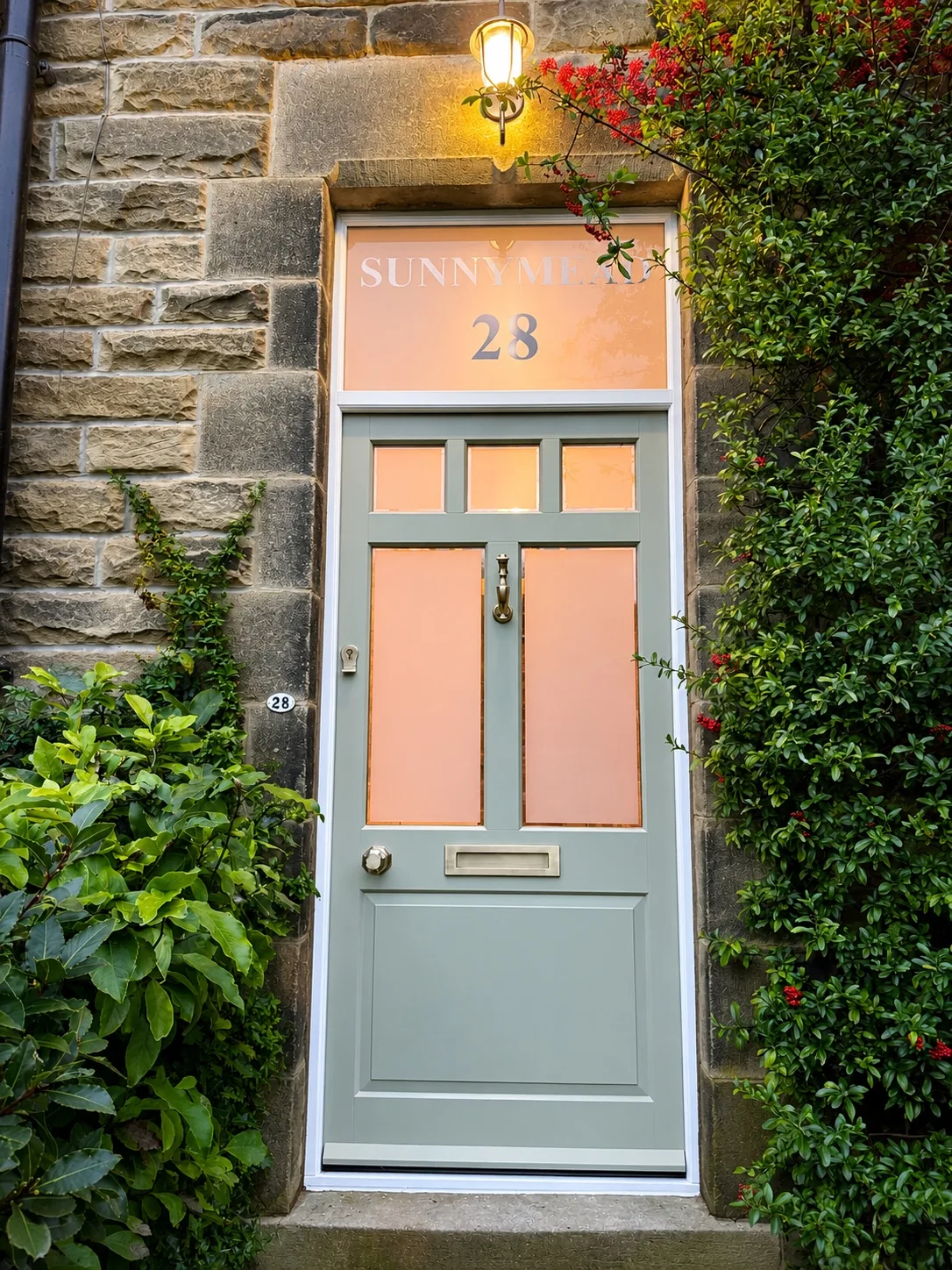 Sage green cottage front door with brass Doctor knocker, brass knob and letterplate, obscure glass panels and etched 'Sunnymead 28' transom, framed by pyracantha and laurel — bespoke heritage door by Bailey Hague Joinery, Yorkshire.